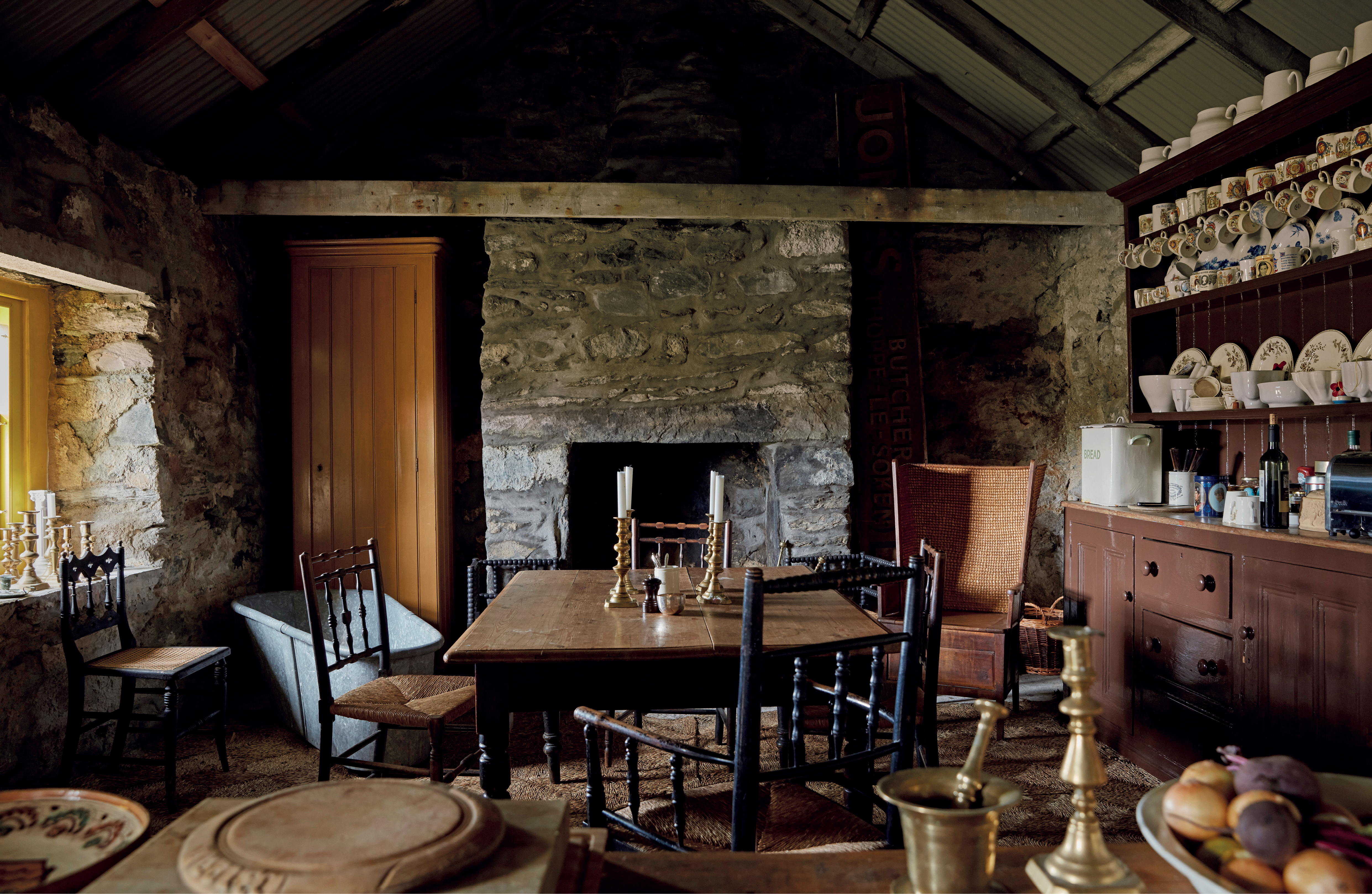 Interior view of a rustic stone cottage kitchen with a wooden table and chairs, a fireplace, and a hutch filled with dishes.